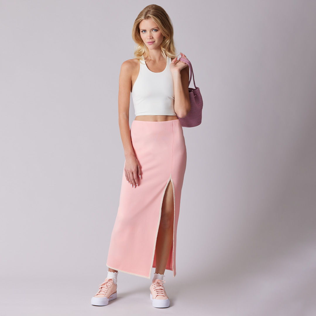 A woman in a white crop top and pink skirt poses against a gray background.