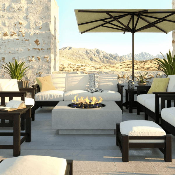 Outdoor lounge seating arranged around a square concrete fire pit on a desert patio, with cushioned club chairs and a market umbrella.
