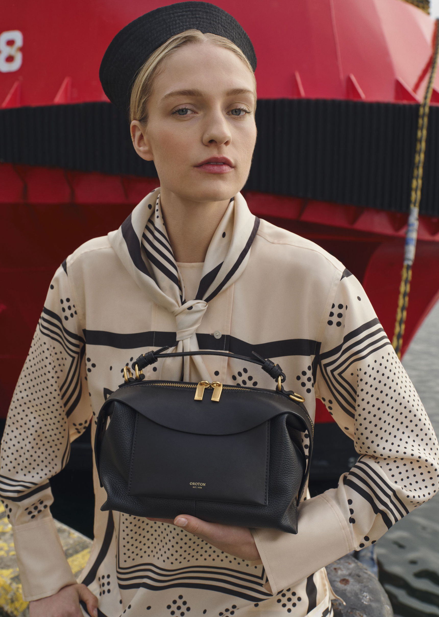Model holding a black handbag, wearing a patterned outfit, with a red background.