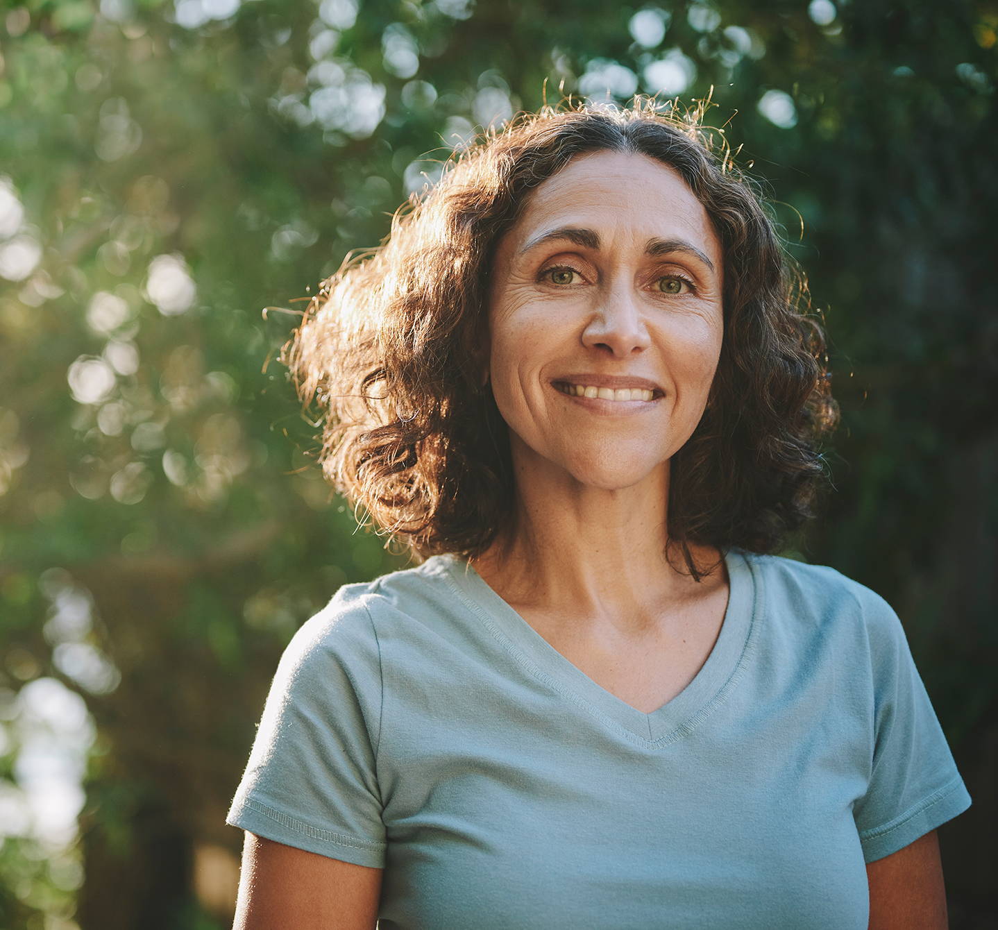 Eine Frau in einem T-Shirt lächelt in die Kamera und genießt einen Tag im Sommer im Freien, während im Hintergrund Sonnenlicht und Grünflächen zu sehen sind.