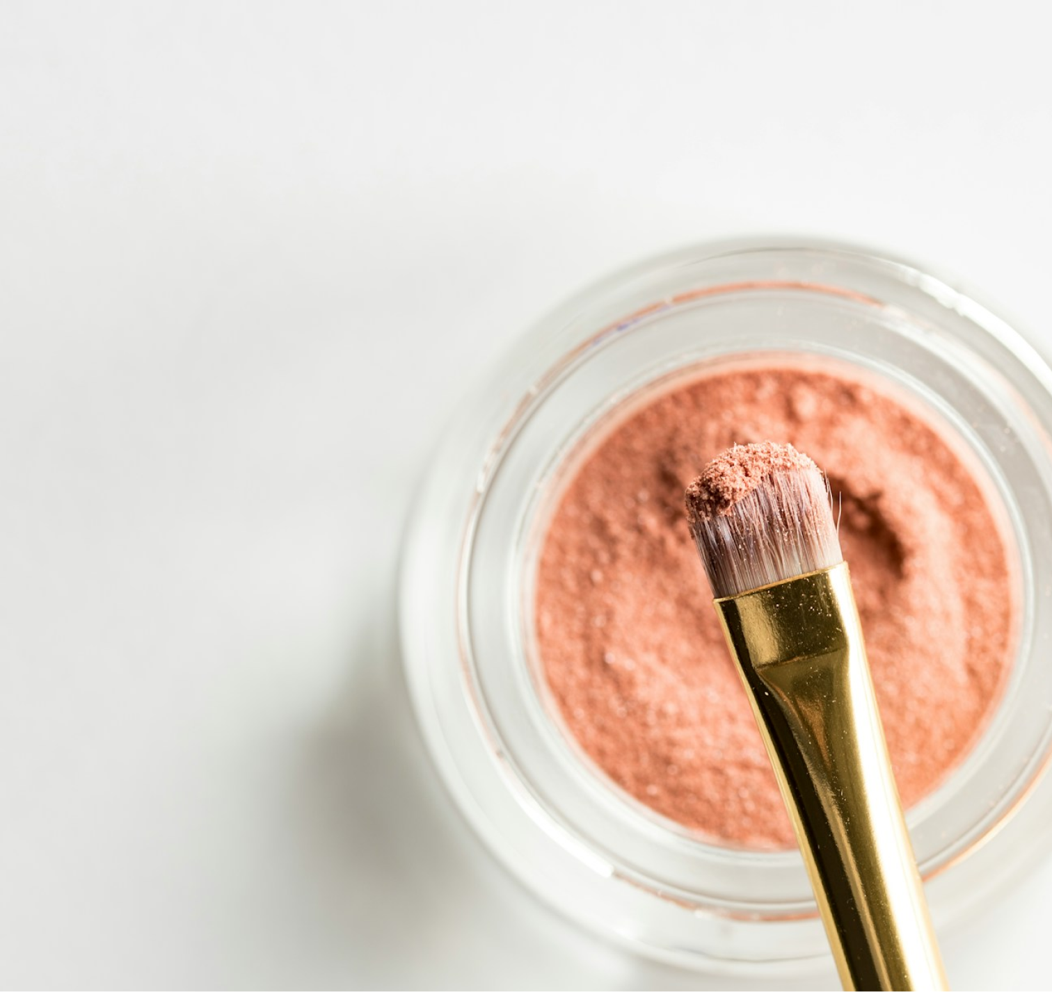 A glass pot containing makeup powder, with a gold-clad brush hovering over it, viewed top down.