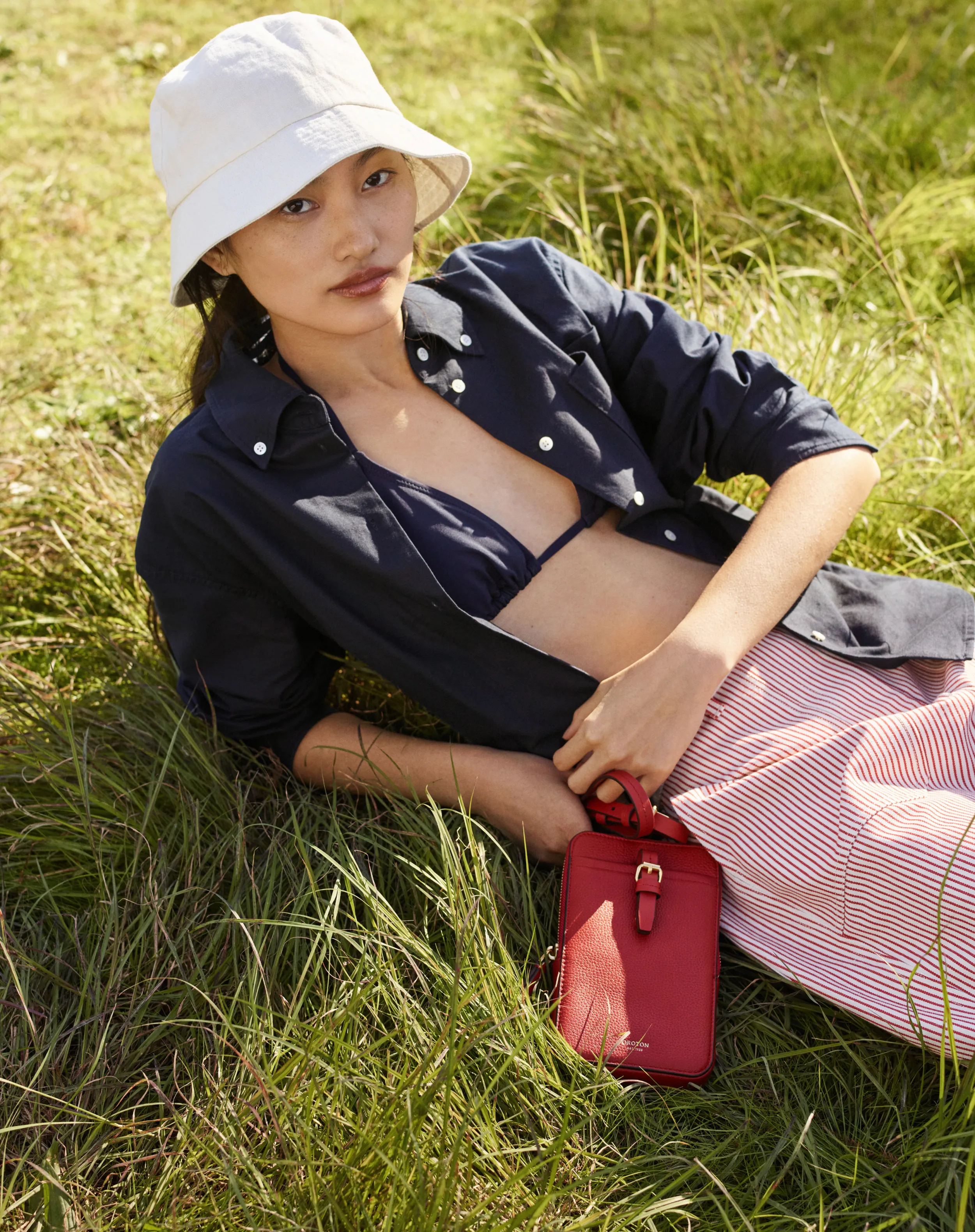 Person in a hat and bikini lying in grass, holding a red bag.