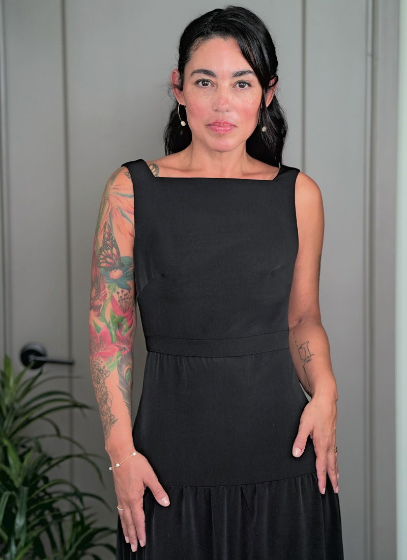 A woman in a black dress stands in front of a gray wall.