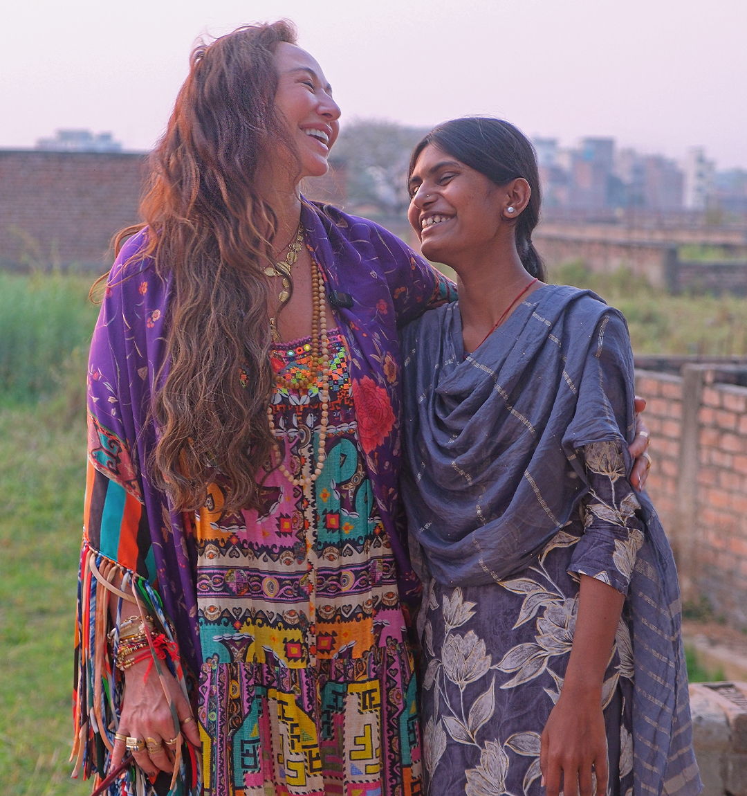 Camilla Franks, Founder and Creative Director, laughs with a girl in a grey-blue scarf, in front of a brick wall and field