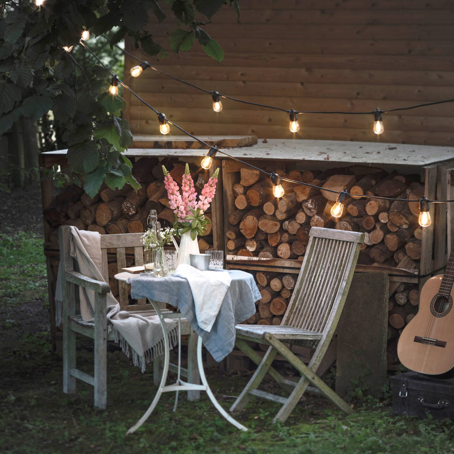 Ultimate Connect festoon lights over an outdoor seating area with logs and a guitar in the background. 
