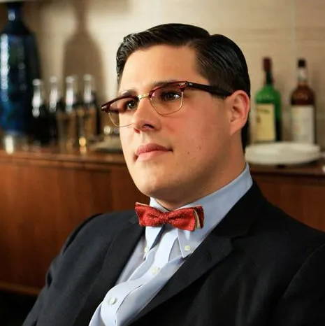 L'acteur Rich Sommer porte des lunettes Browline écaille de tortue avec un nœud papillon rouge et un costume bleu marine foncé dans la série télévisée Mad Men