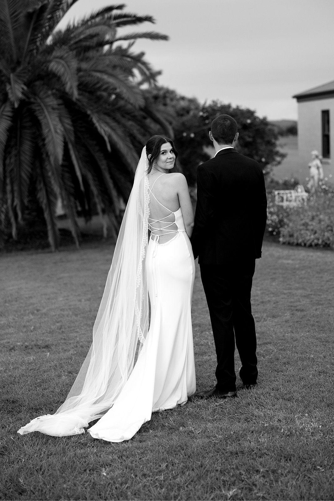 Bride and groom standing together outdoors, facing away, with a palm tree nearby.