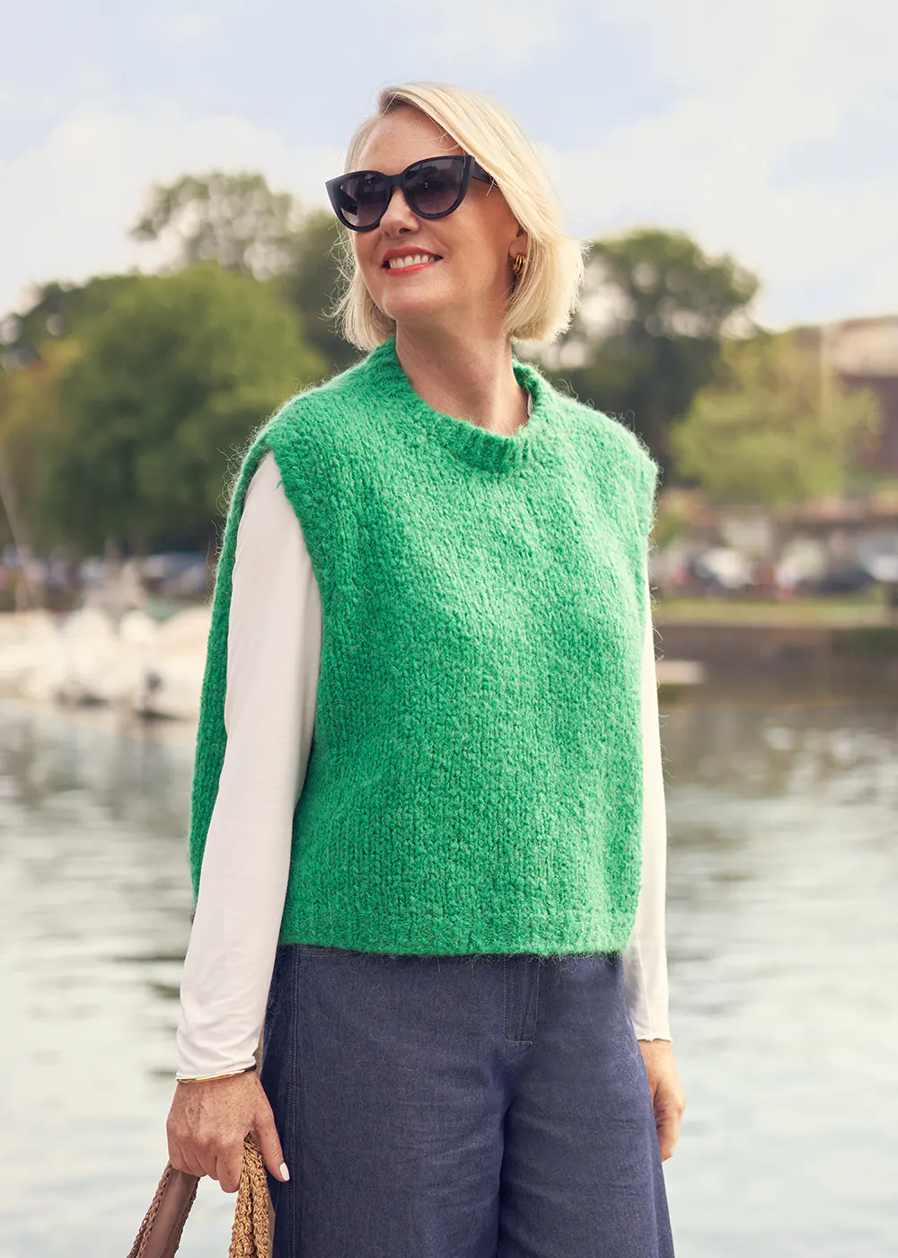 Woman wearing a green sleeveless sweater over a long sleeve white tee and blue trousers