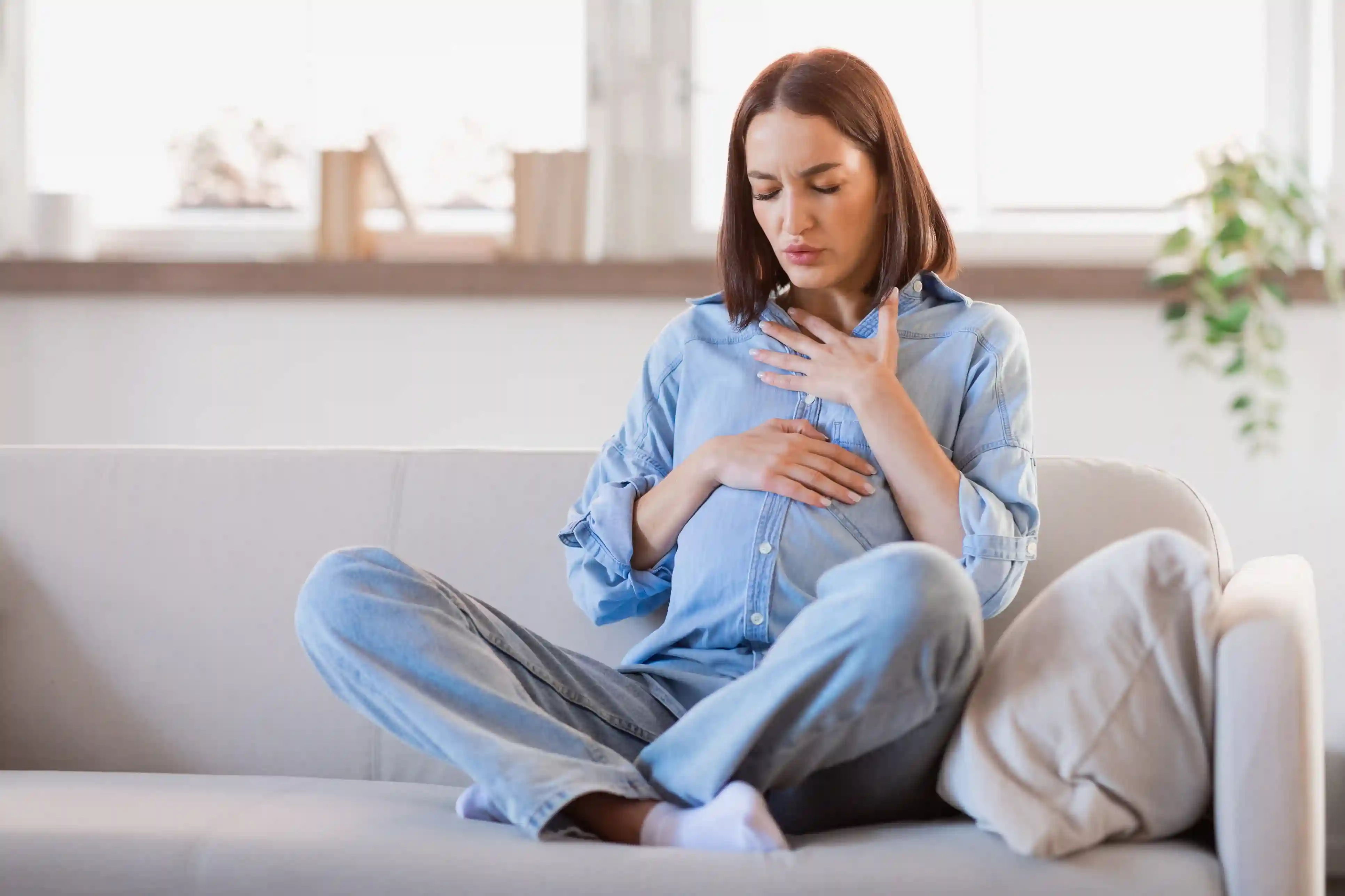En gravid kvinne sitter i sofaen med en hånd på brystet og den andre på magen – astma kan påvirke barnet i magen