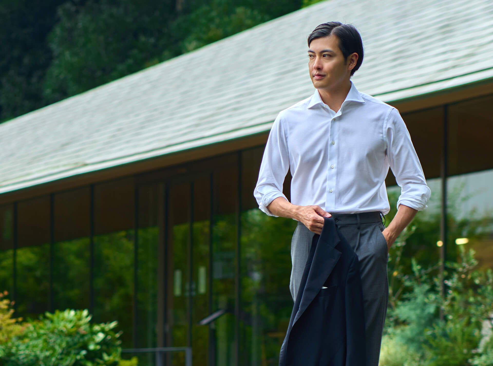 Man in a white shirt holding a jacket outdoors near a modern building.