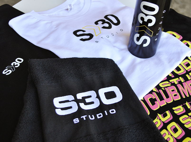 A merch bundle for S30 studio featuring white and black screenprinted t-shirts with an embroidered towel and screen printed drink bottle sitting on top
