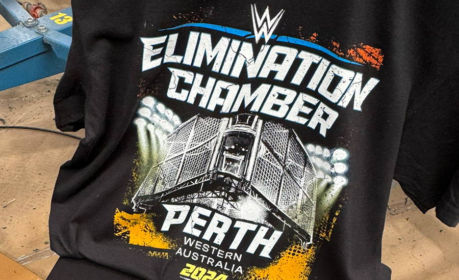 A simulation process screen print on a black t-shirt for the 2024 'WWE Elimination Chamber' event merchandise, behind held up in the factory with a small section of the screenp printing machine arm visible indicating the shirt has just come off the press. The print shows the WWE logo with a bright blue detail line above bold Elimination Chamber white text with a subtle blue keyline, and a heavily distressed grunge style white graphic of the fighting cage and scaffolding above the words Perth Western Australia in white and blue keyline, and 2024 printed in bright yellow below. Either side of the cage is stadium lighting depicted as solid white centres with a yellow and blue outer glow effect and spotlighting created with layered halftone graduation of colour. Sections of orange grunge texture colour highlights have been added at the base and top of the graphic. 