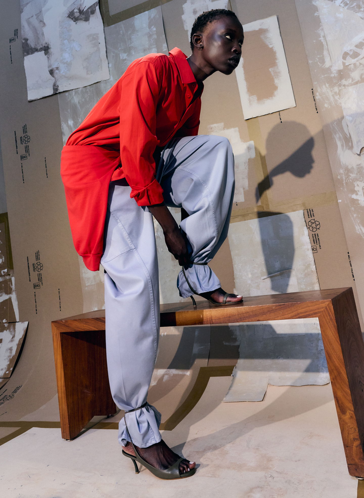 Model wearing a red top and gray pants poses on a wooden bench.