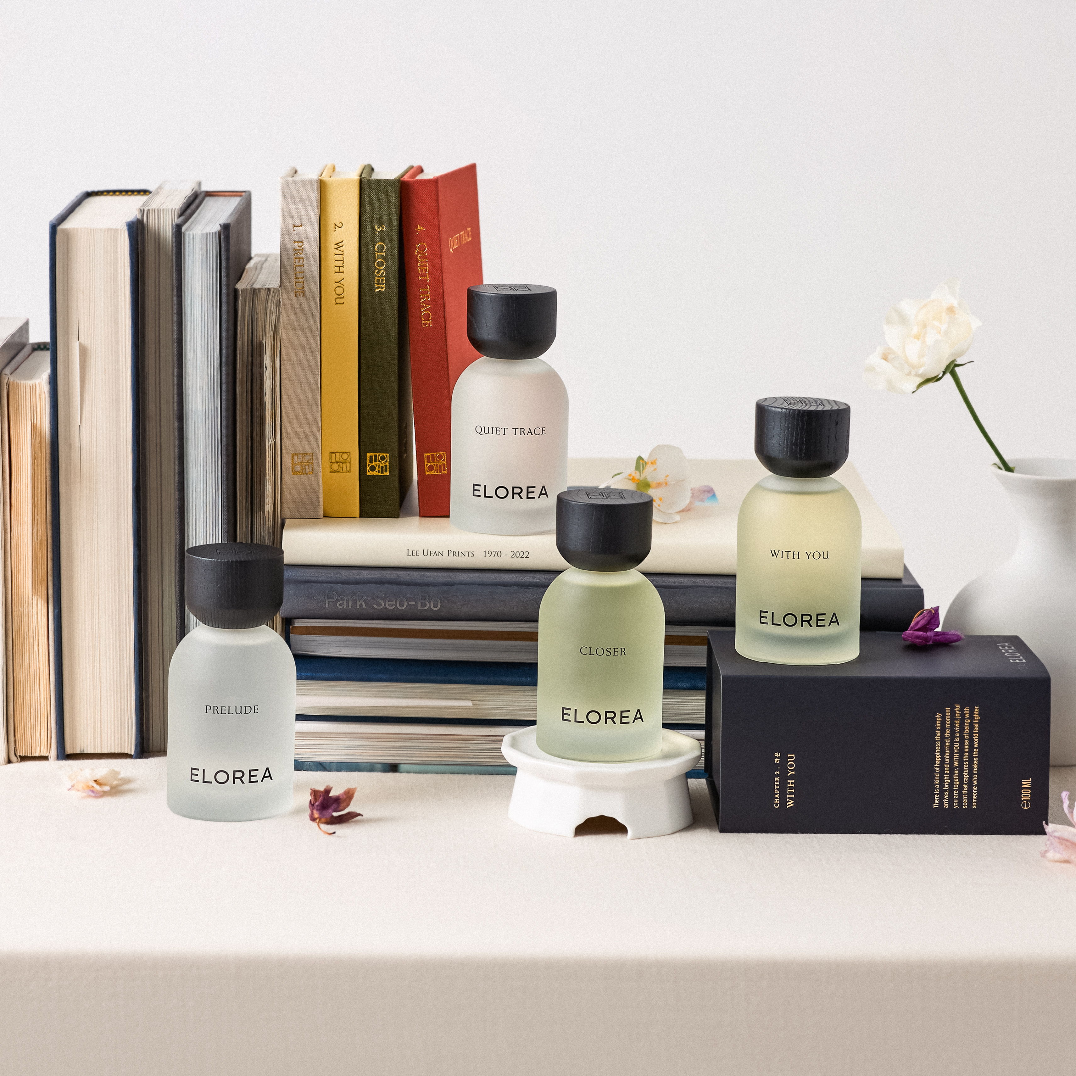 Bottles of fragrance on a table with stacked books and a flower vase.