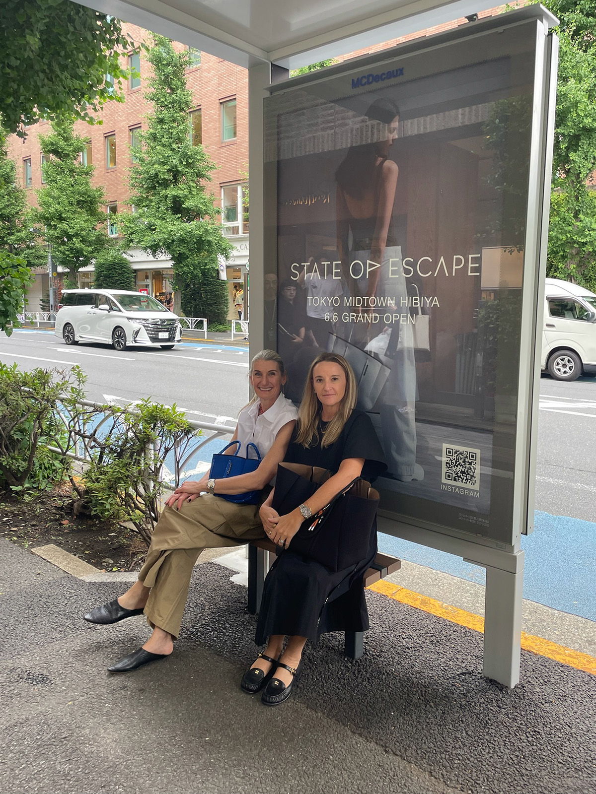 Two women sitting at a bus stop with an advertisement behind them.