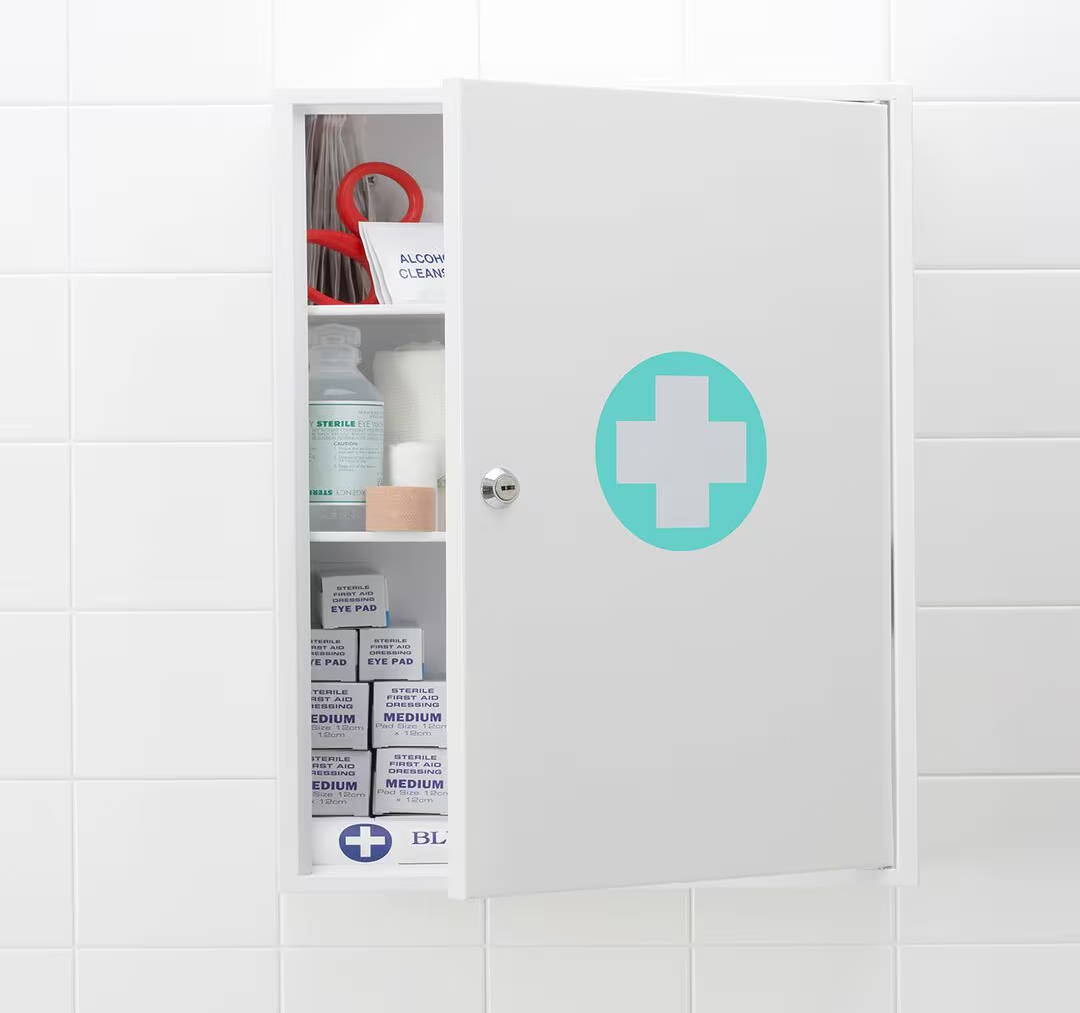Skrinka v kúpeľni s polootvorenými dverami, na ktorých sa nachádza biely kríž v zelenom kruhu, symbolizujúci úložný priestor pre lieky
