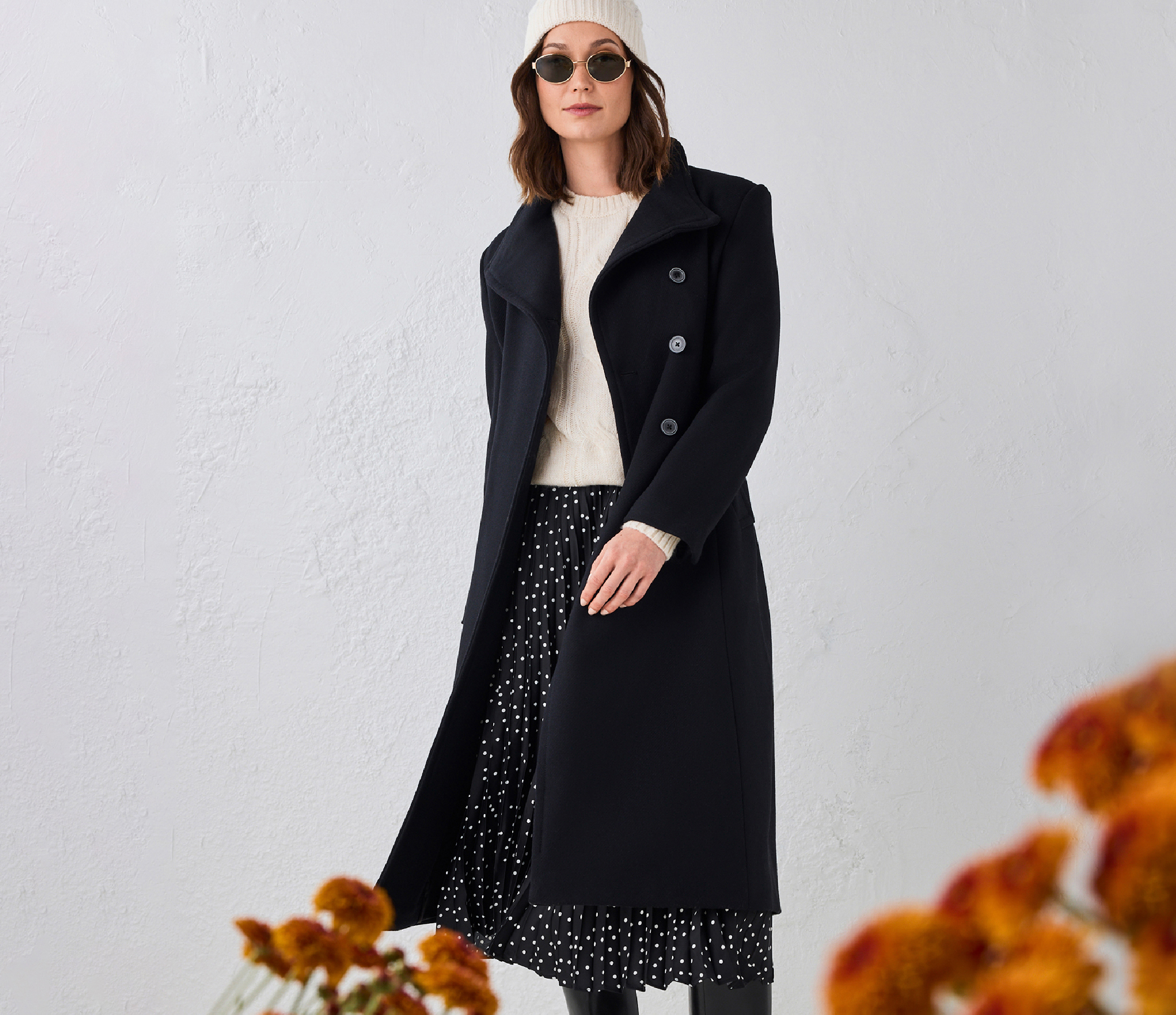 Woman in a black coat and sunglasses, standing against a light background.