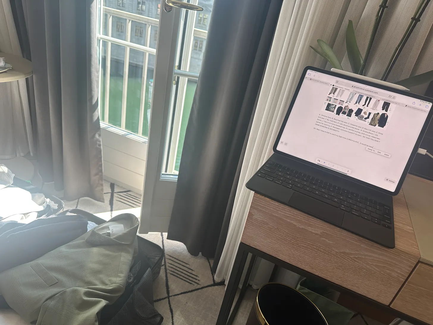 A laptop on a desk near a window with a suitcase nearby.
