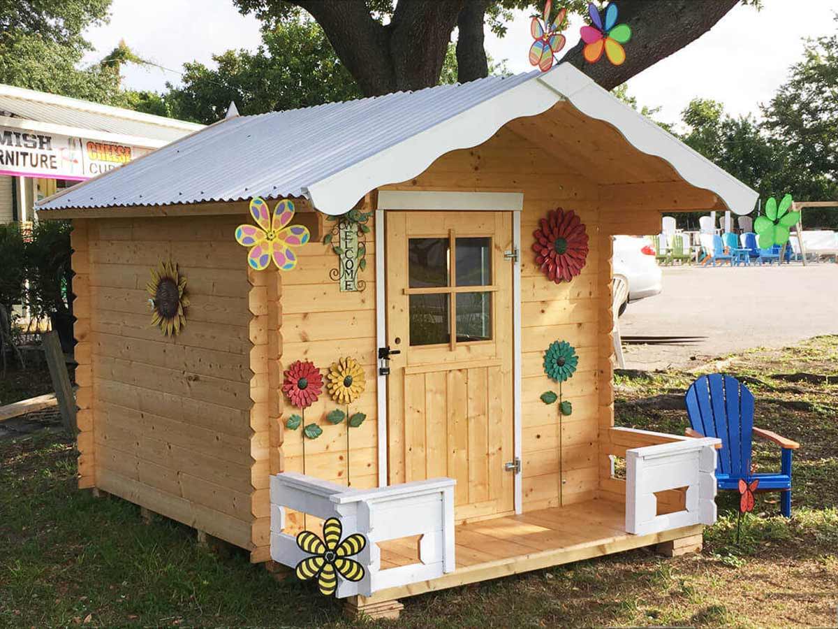 Wooden DIY playhouse with a white roof and decorative flowers with a blue chair next to it by WholeWoodPlayhouses