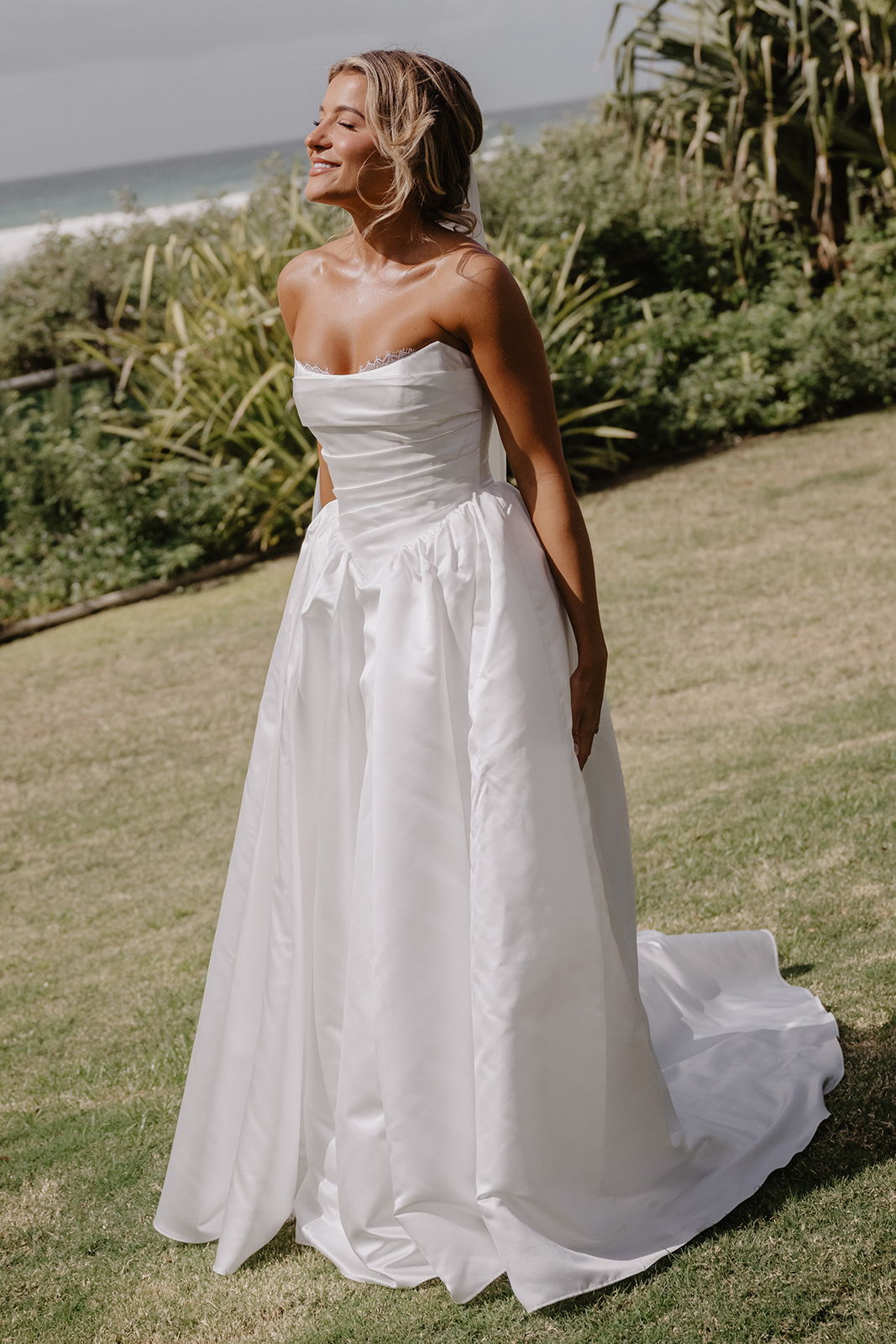 Woman in a white wedding dress standing on grassy outdoor area.