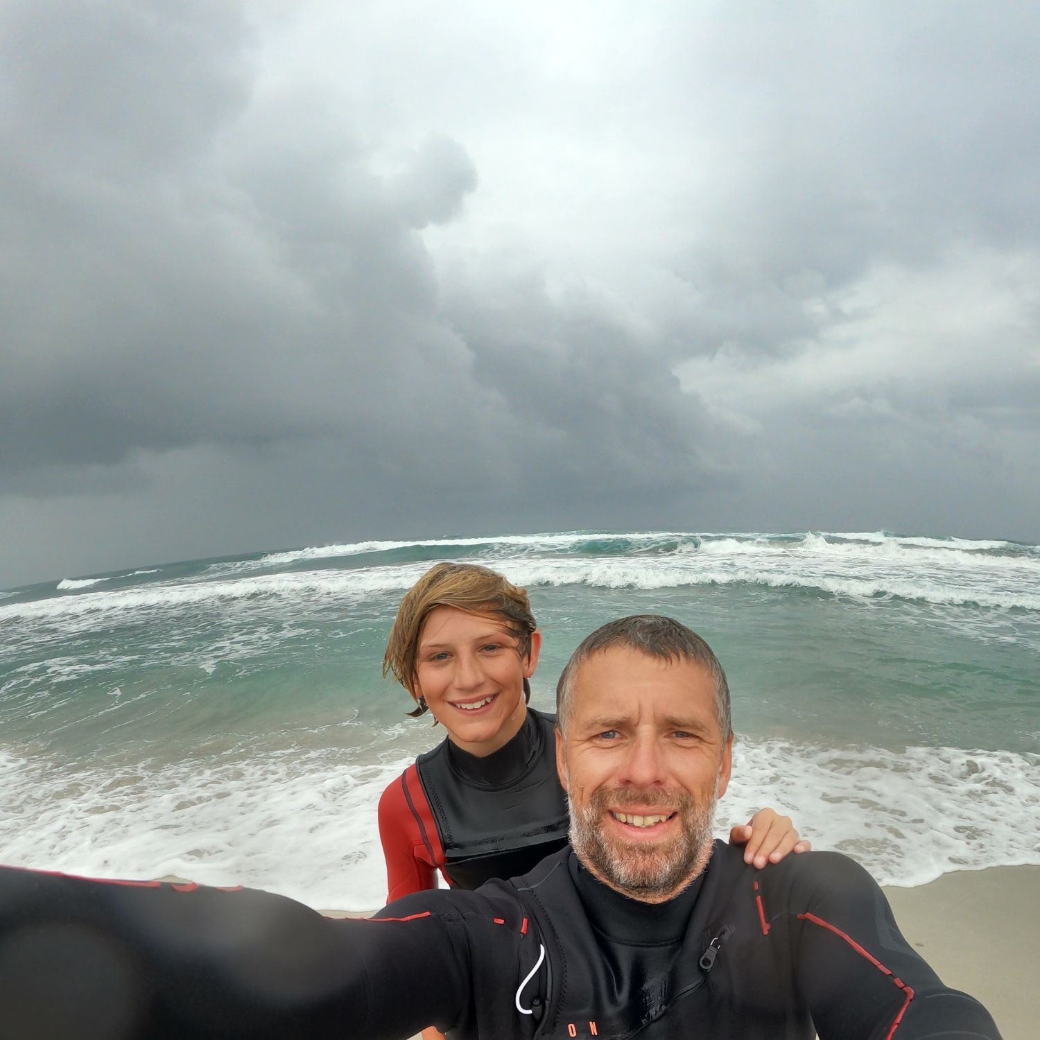 A couple in wetsuits taking a selfie at the beach with waves.