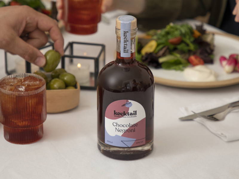 Chocolate negroni cocktail bottle on dinner table with people picking up olives in background