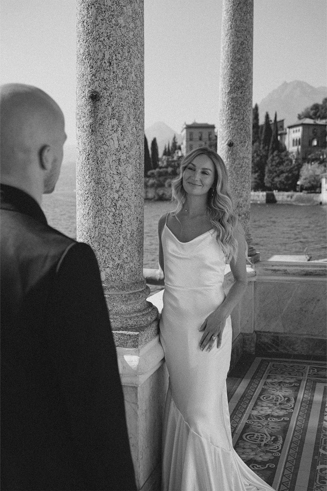 Bride in elegant dress smiles at groom by scenic waterfront.