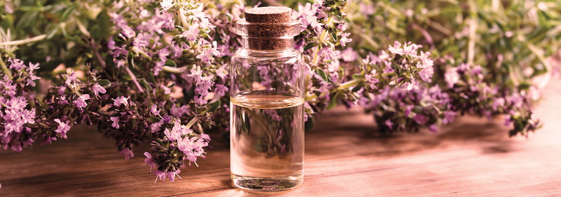 Glass bottle of oil surrounded by purple flowers on a wooden surface.
