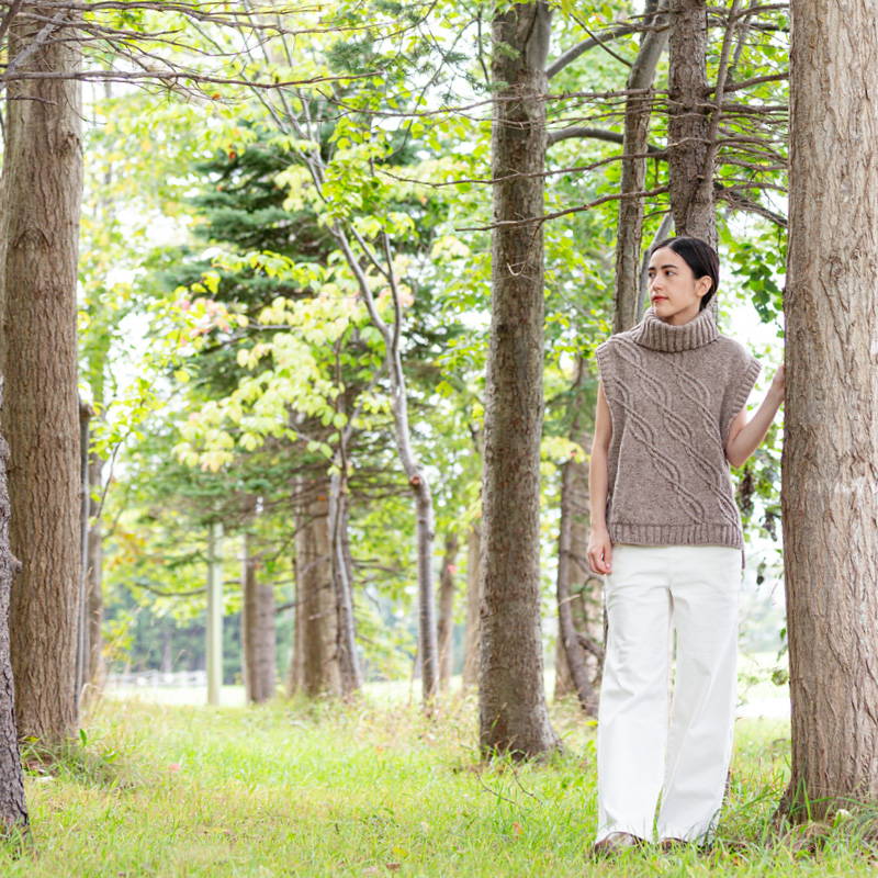 A woman in a wooded area leans against a tree and is modeling Gyration turtleneck vest by Keiko Kikuno in Brooklyn Tweed Shelter