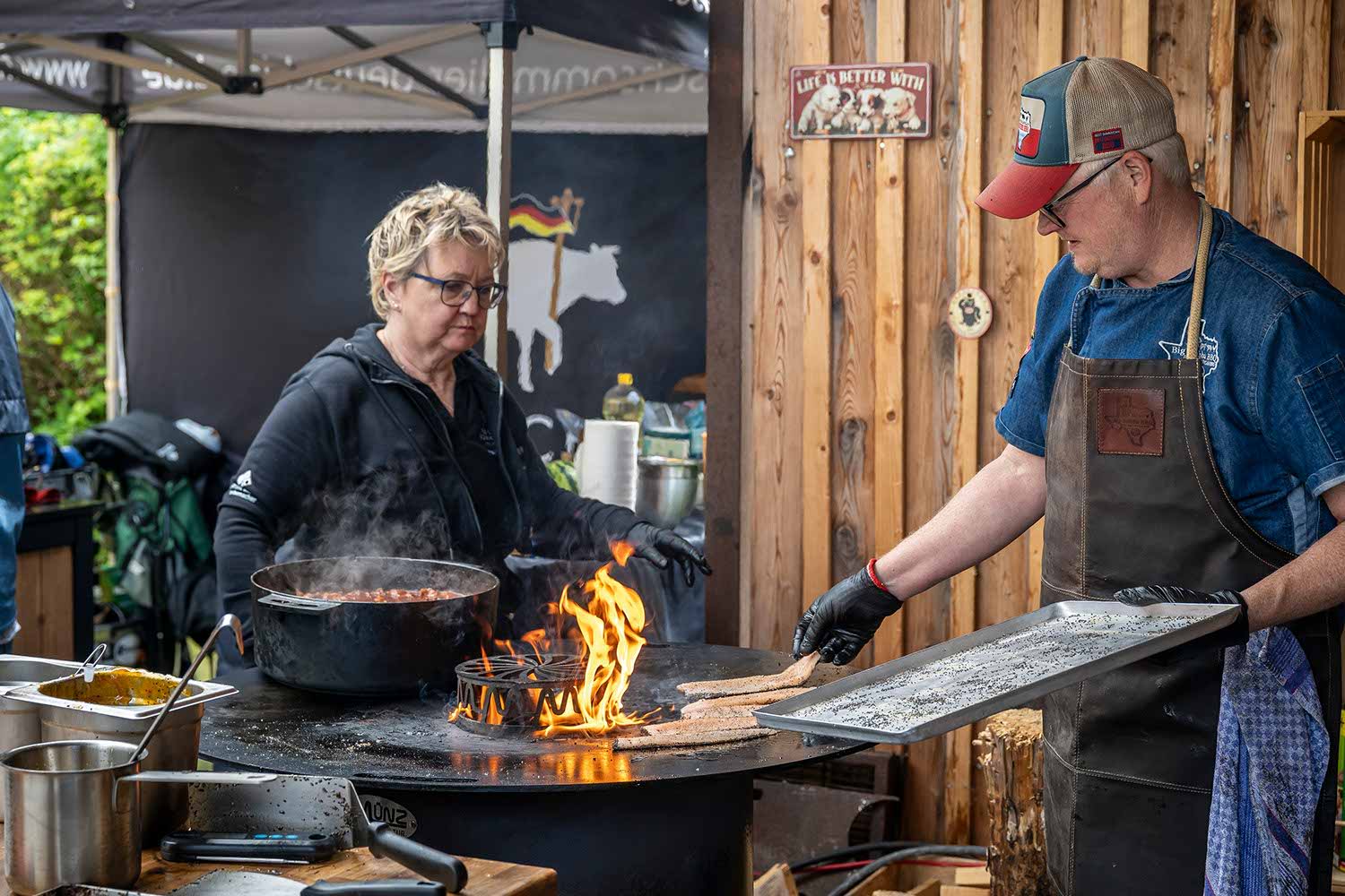 Two cooks preparing food with flames in an outdoor kitchen setting.