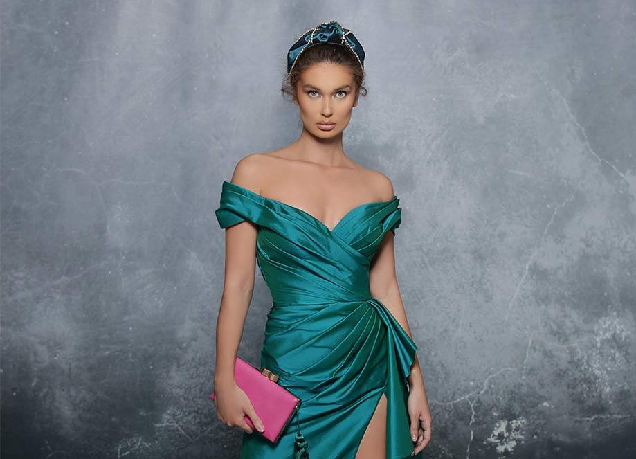 Woman in emerald off-shoulder gown with pink clutch and tiara poses against textured grey background.