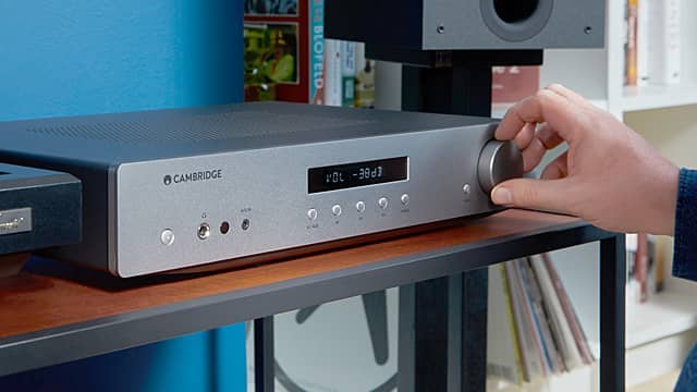 A person adjusting a silver audio receiver on a wooden shelf.