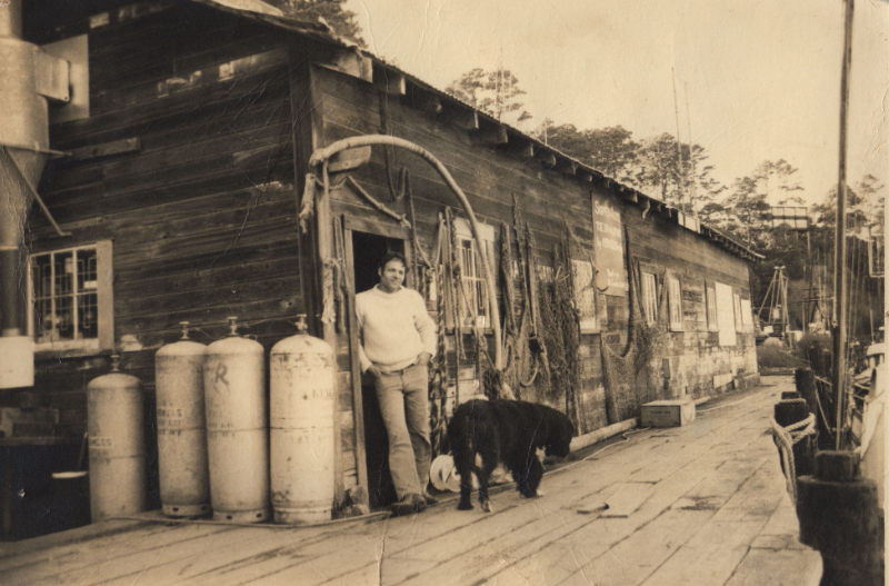 A man stands outside a wooden building with a dog nearby.
