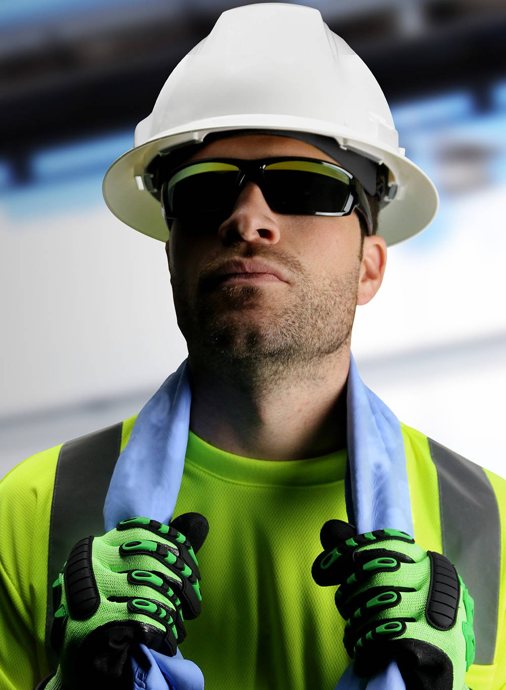 Construction worker wearing Hi-viz lime safety shirt, and white hard hat holding blue cooling towel around neck with dark safety glasses. He is also wearing green impact resistant gloves.