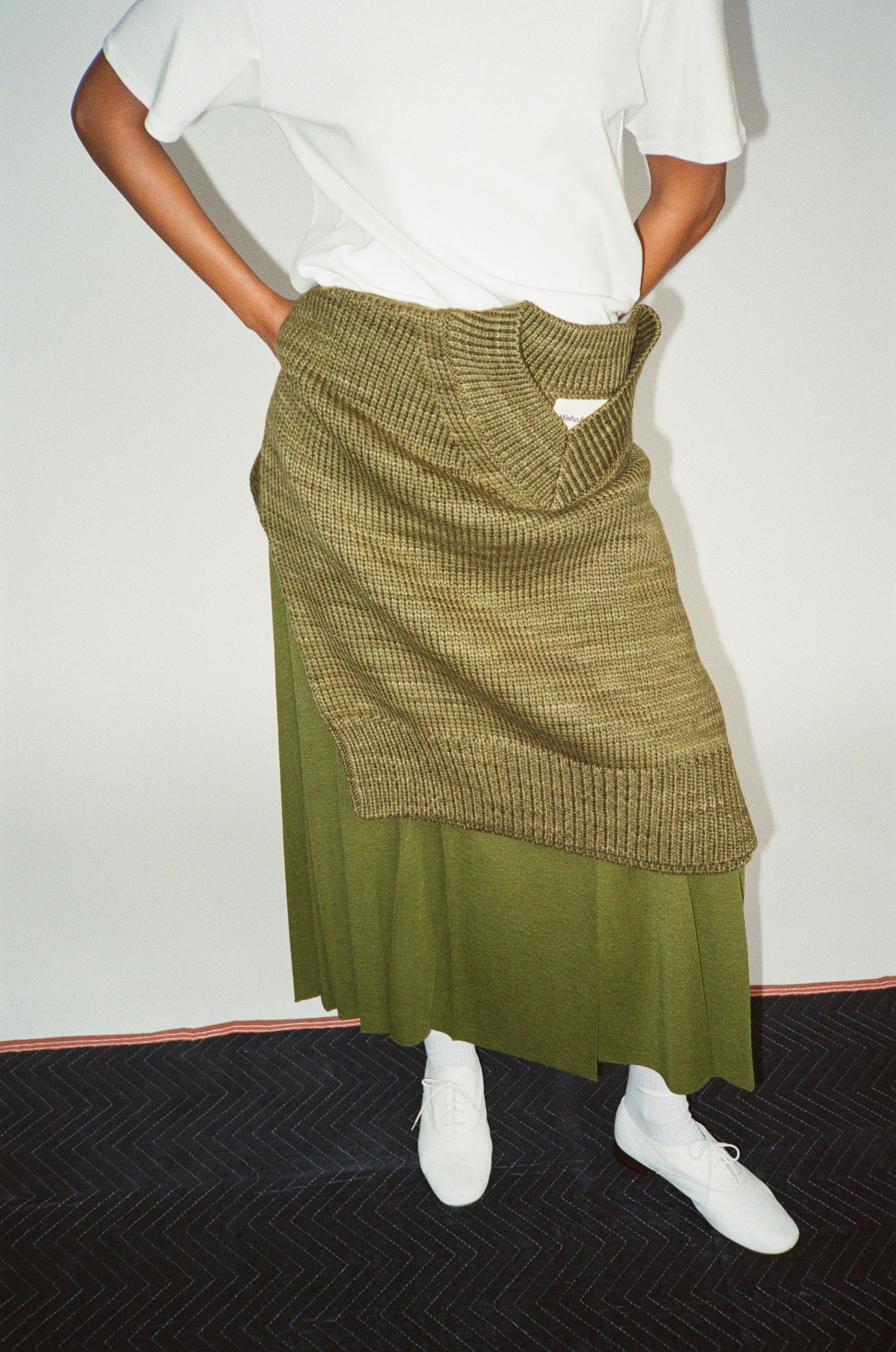 Person wearing a white shirt, green skirt, and white shoes, posing indoors.
