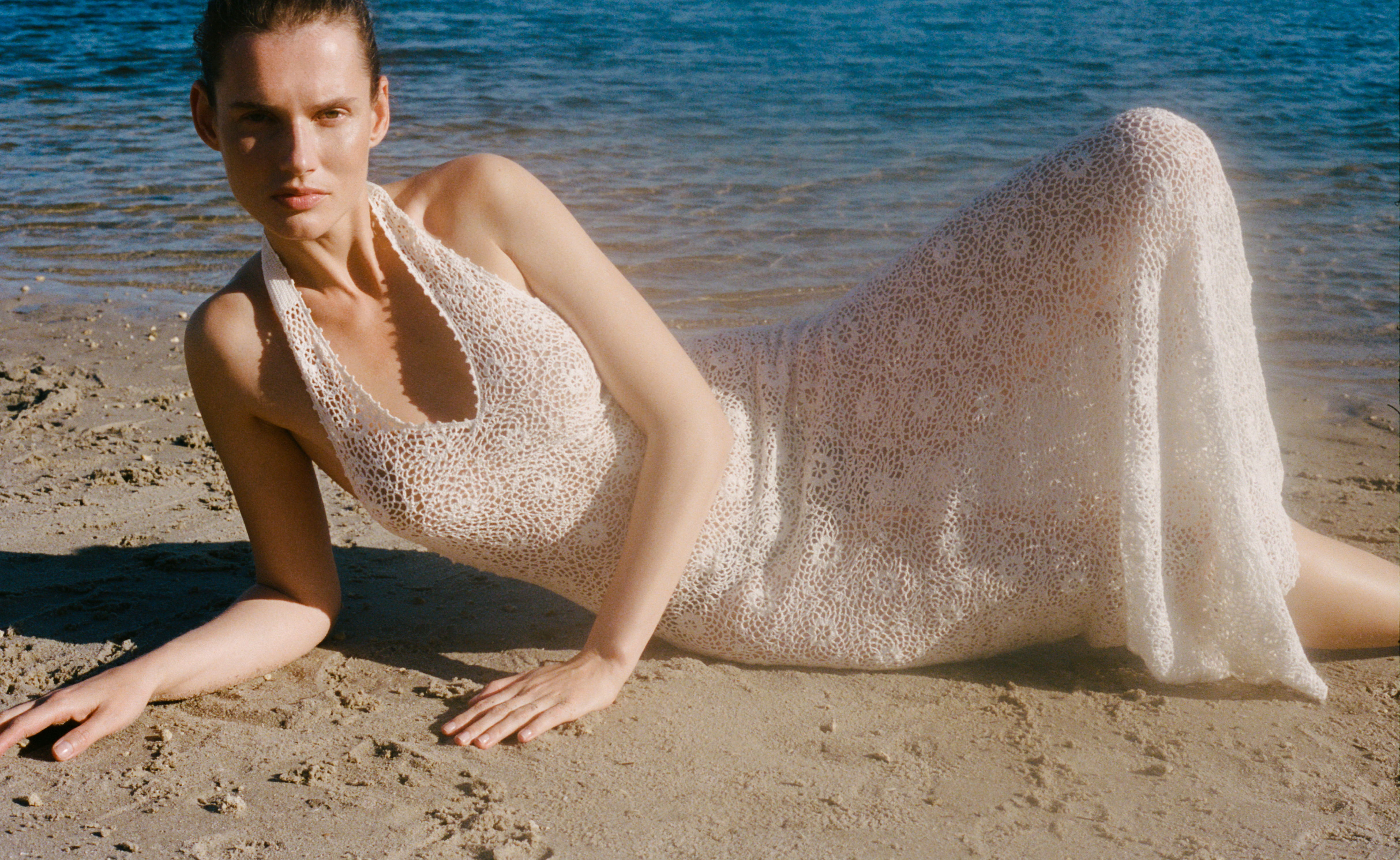 A woman in a white dress lounging on the beach by the water.