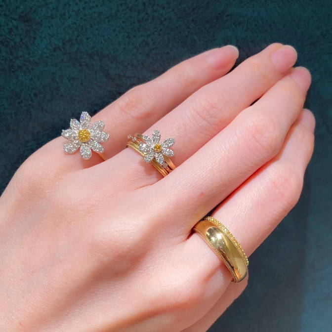 A hand wearing two flower rings and a gold band.