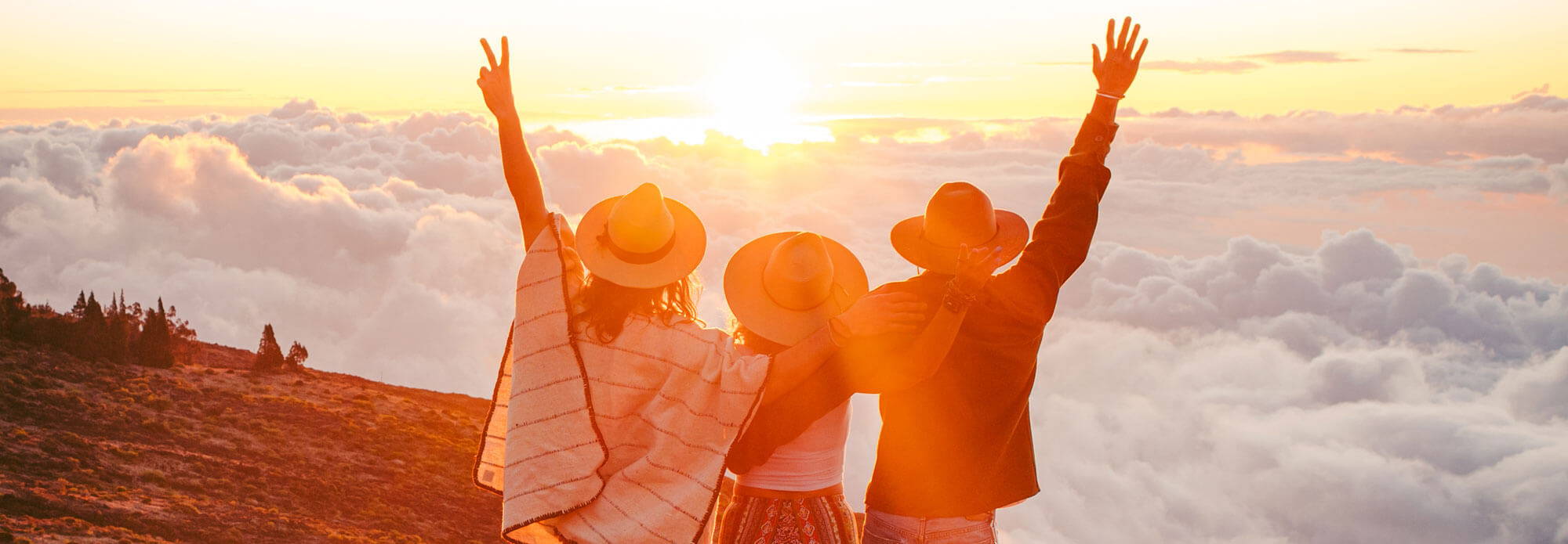 Three friends wearing sun hats standing on a cliff during sunset