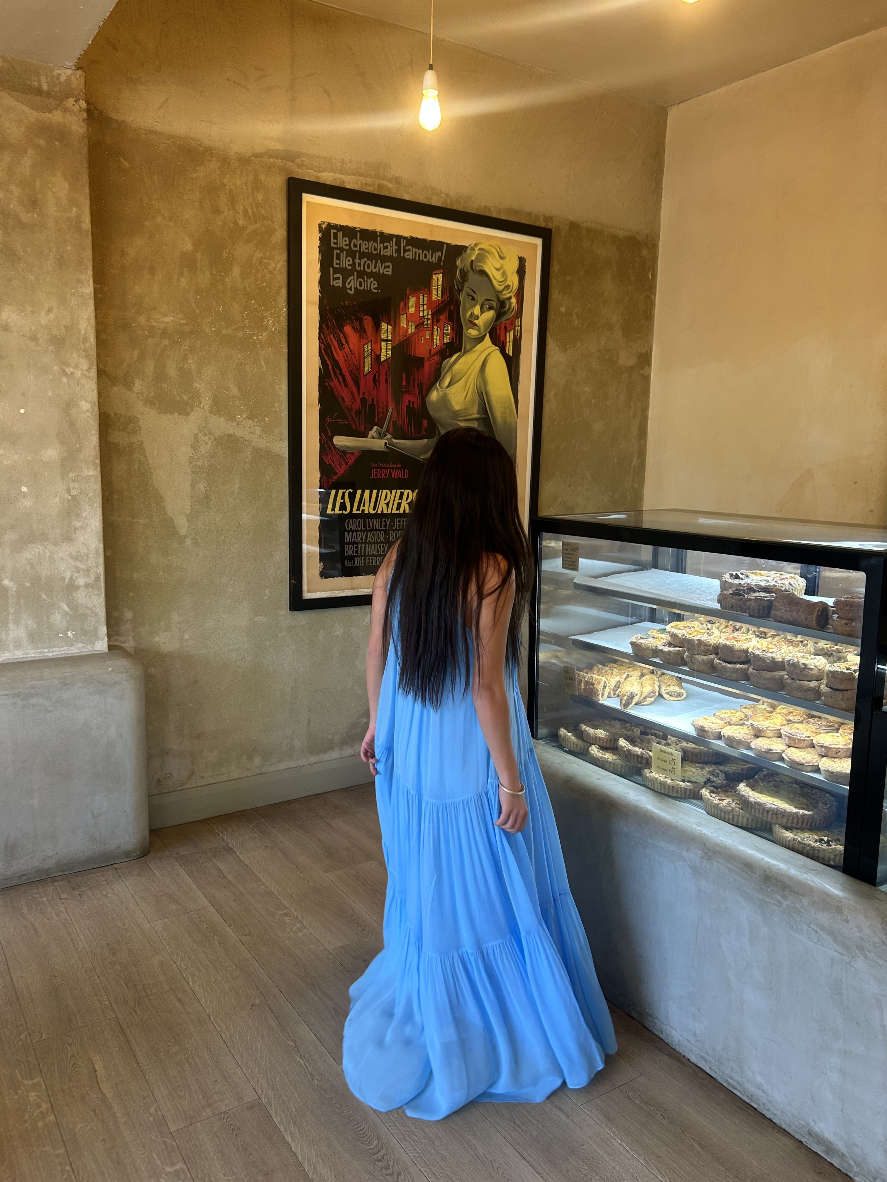 Frankie stands in labanette looking at the pastry cabinet. She is wearing the Jena Cami Dress in Sky.