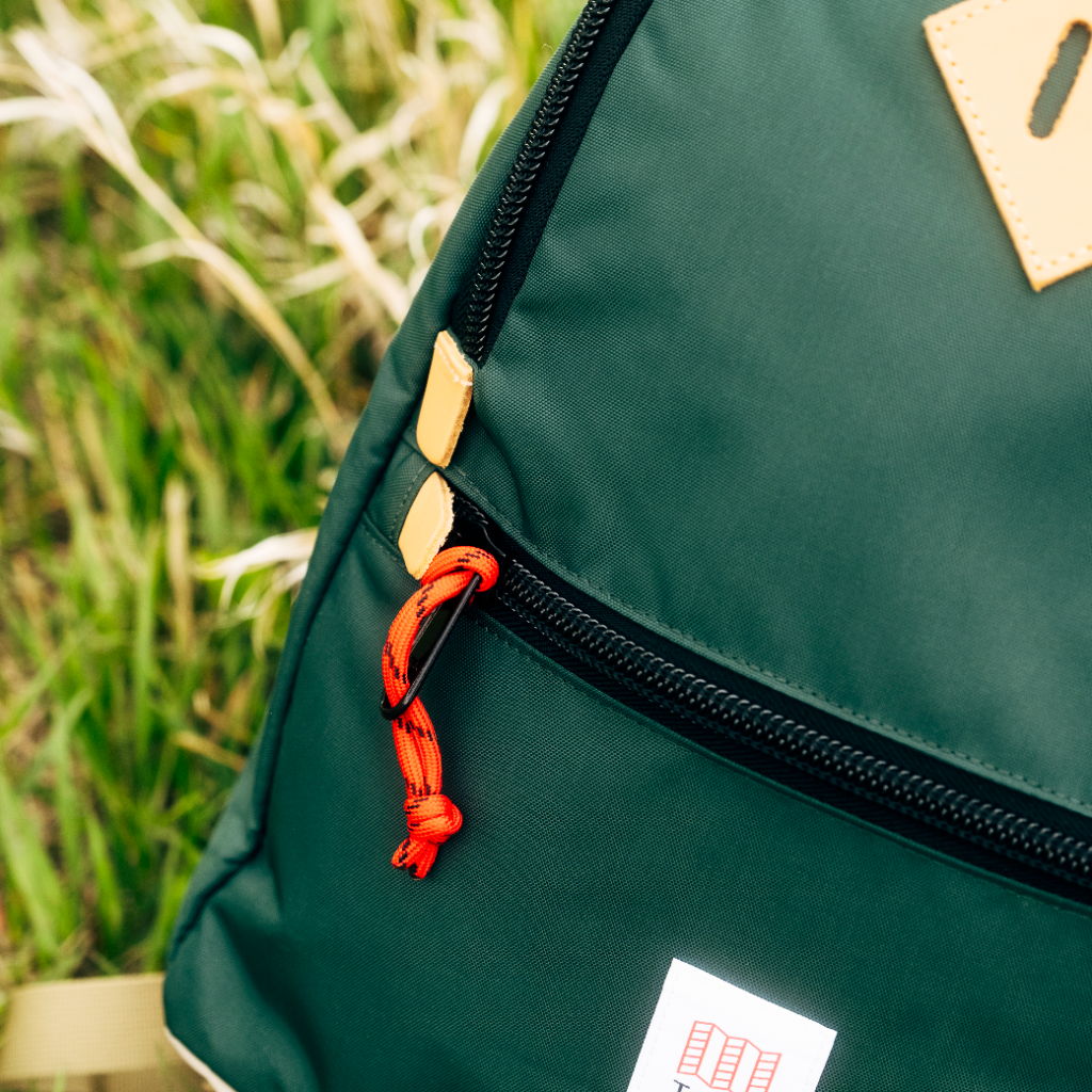 Green backpack with a zipper and orange pull tab, set against grass.