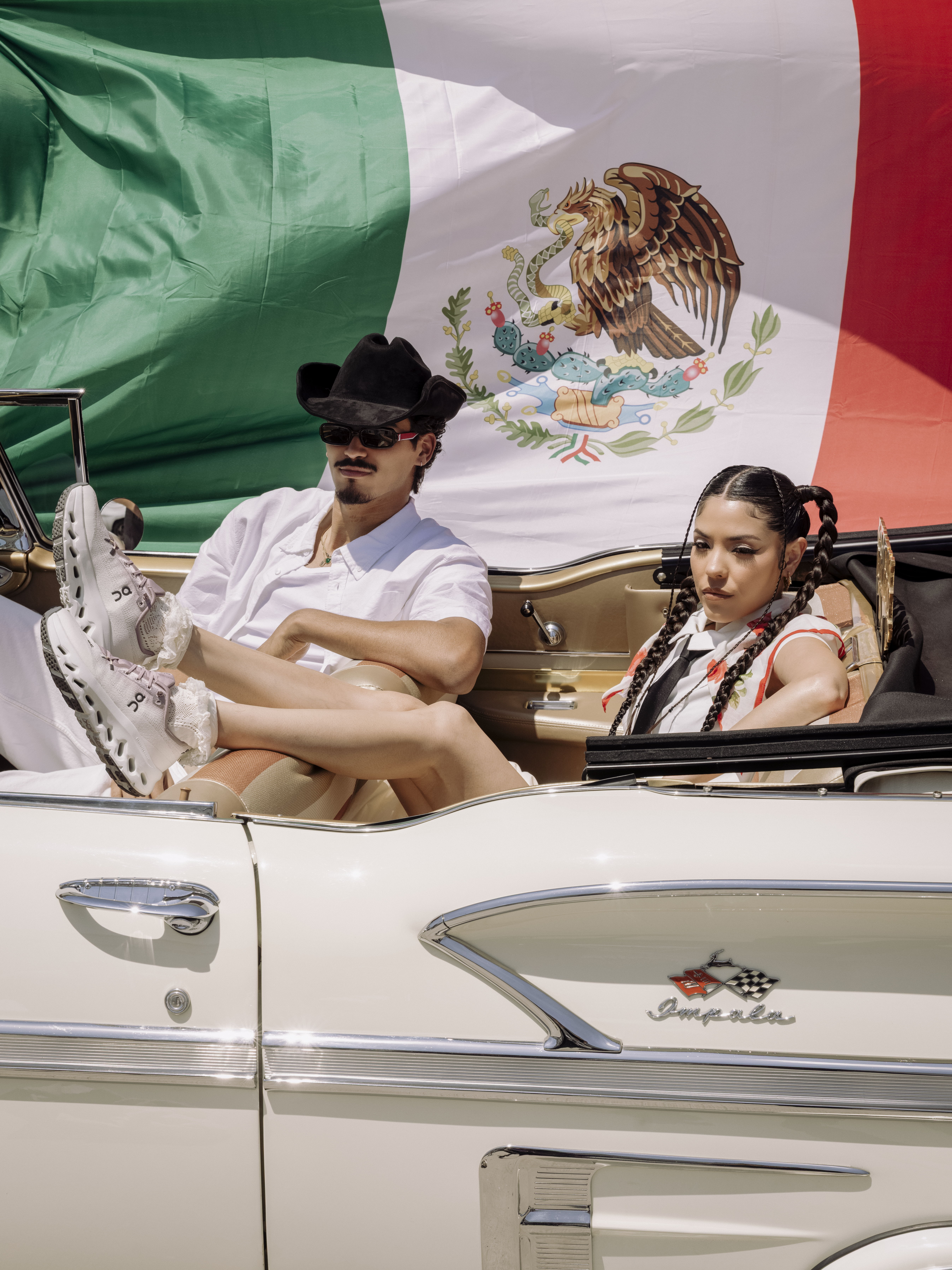 Man and woman in vintage car, with a Mexican flag backdrop.