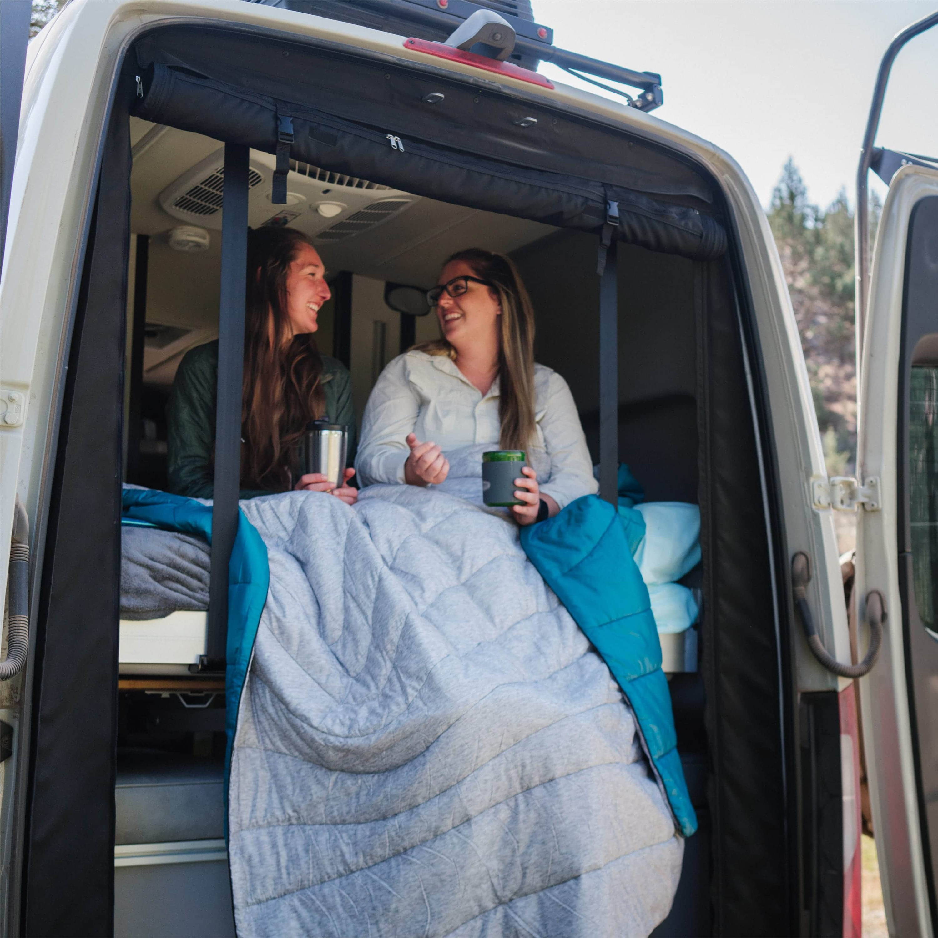 Two Women Drinking Coffee Outside Of Back Of Car While Wrapped In an Original Puffy Blanket