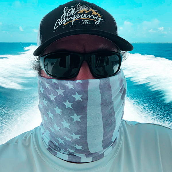Jason Gascoyne on a boat with the wakes in the background, looking into the camera while wearing a face shield over his face, sunglasses and a hat.