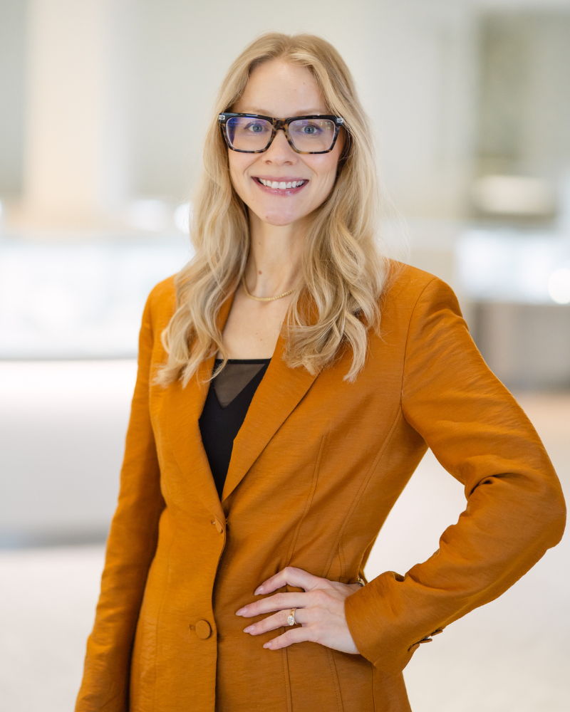 Smiling woman in an orange blazer with glasses in a bright setting.