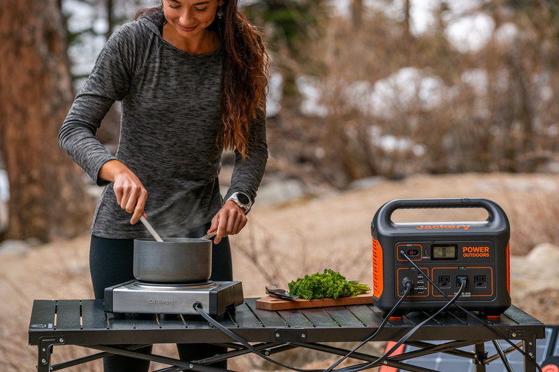 A smiling woman uses a portable solar power station to cook a meal in the woods.
