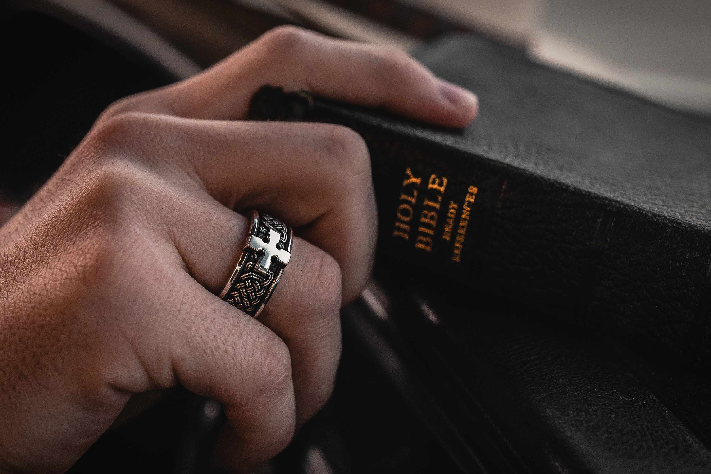 The Hand of a Man Wearing the Joshua Cross Band Ring and Reaching for a Bible