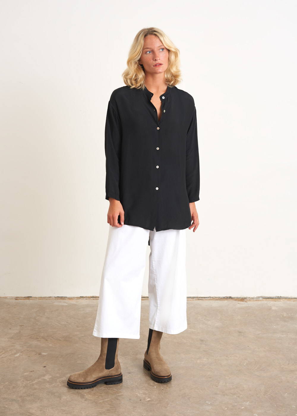 A model wearing a black oversized shirt with mother of pearl buttons over white culottes and taupe chelsea boots