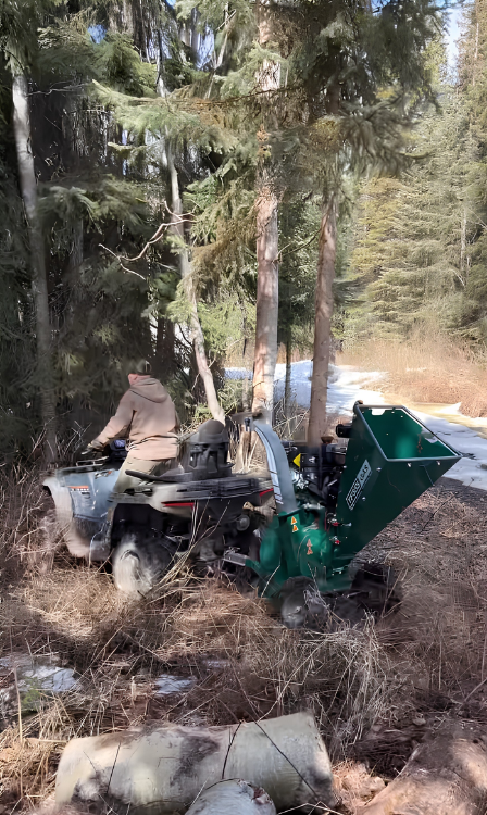 Person using a wood chipper attached to an ATV in a forested area.