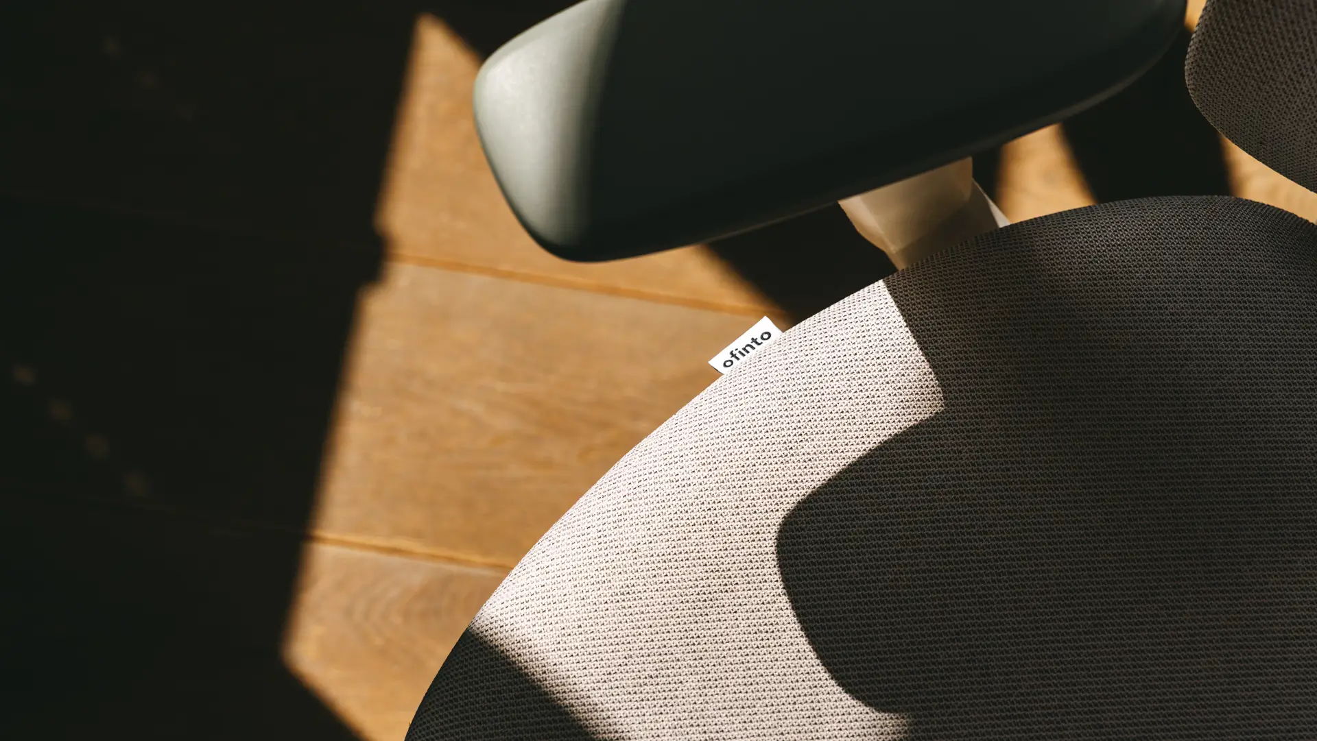 Gros plan sur l'assise claire d'une chaise de bureau portant le label « ofinto », avec un accoudoir noir et une base en bois ; jeu d'ombres et de lumières dans l'image.