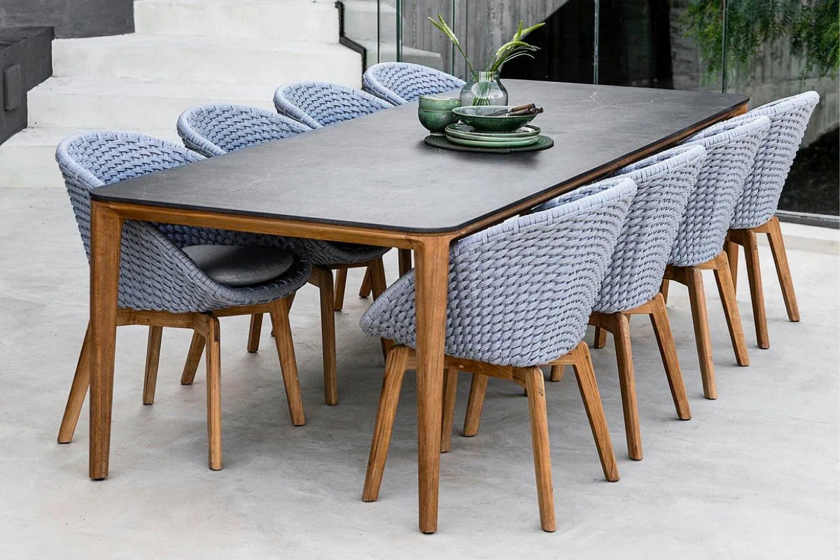 An outdoor table with smooth teak legs and stone top accompanied by 8 woven light grey dining chairs with matching teak legs.